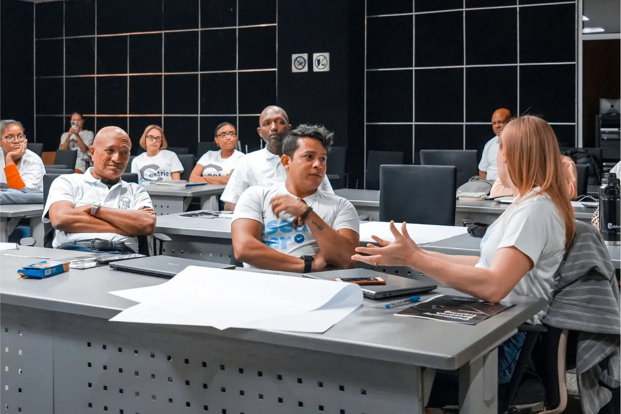 Community researchers in white Centric T-shirts sit at desks during a training session in the Dominican Republic, while one participant speaks.