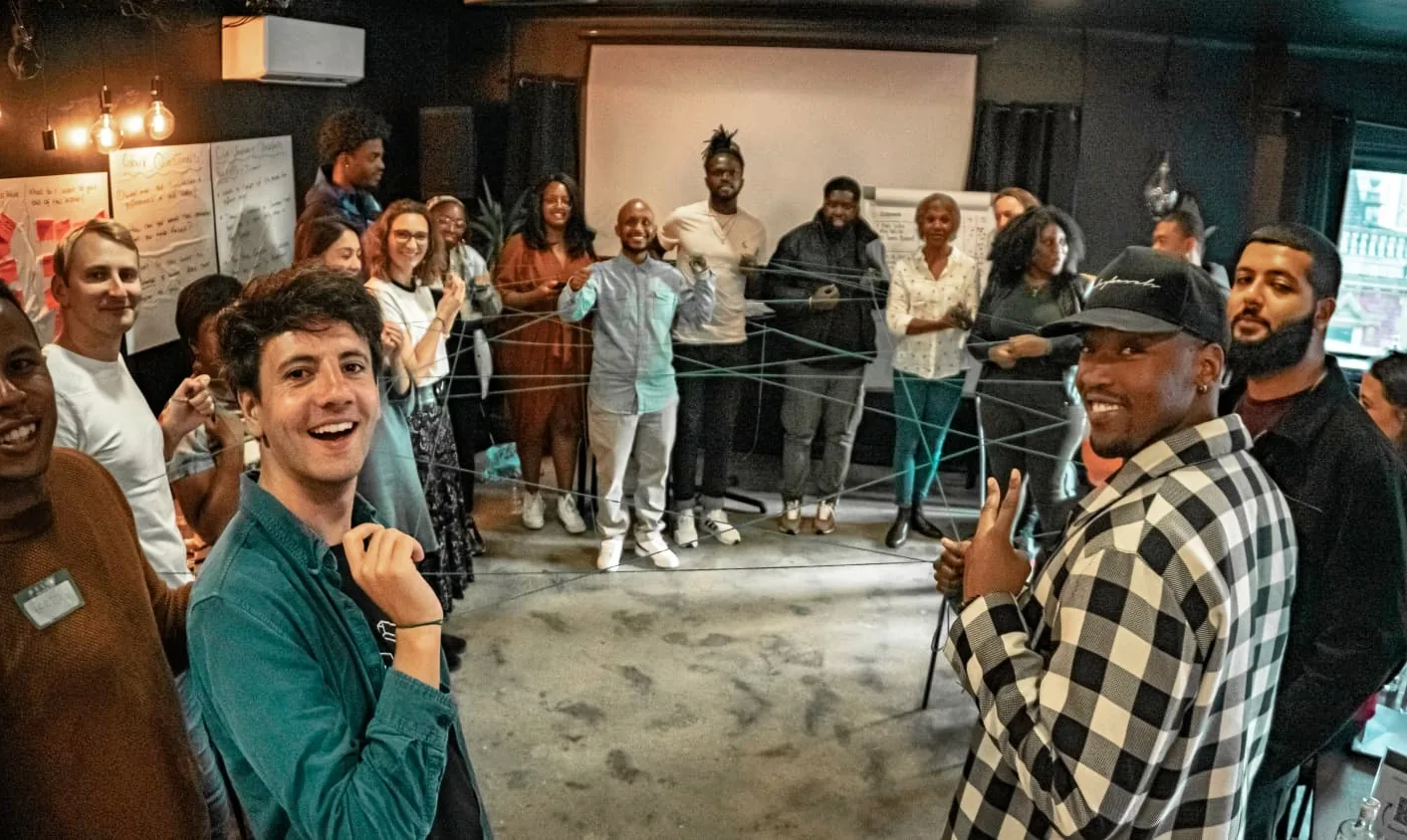 Around twenty people stand in a circle at a race and reflexivity workshop, holding a web of string stretched between them, smiling at the camera.
